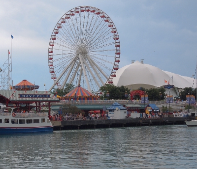 [Large Ferris wheel, white domed building, circus-colored tents on ground and ships lining the pier.]